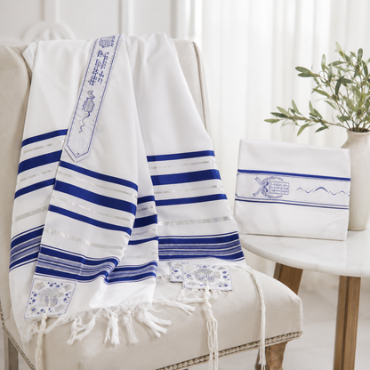 A white Jewish tallit prayer shawl with blue stripes and embroidered details draped over a beige armchair, with a matching bag set on a side table in a bright, softly lit room.