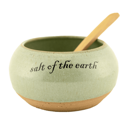 Seafoam green ceramic salt bowl that  says "salt of the earth" in English and Hebrew with a wooden spoon