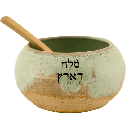 Seafoam green ceramic salt bowl that  says "salt of the earth" in English and Hebrew with a wooden spoon