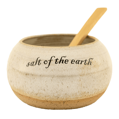 Pearl-colored ceramic salt bowl that says "salt of the earth" in English and Hebrew with a wooden spoon