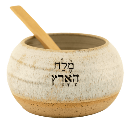 Pearl-colored ceramic salt bowl that says "salt of the earth" in English and Hebrew with a wooden spoon