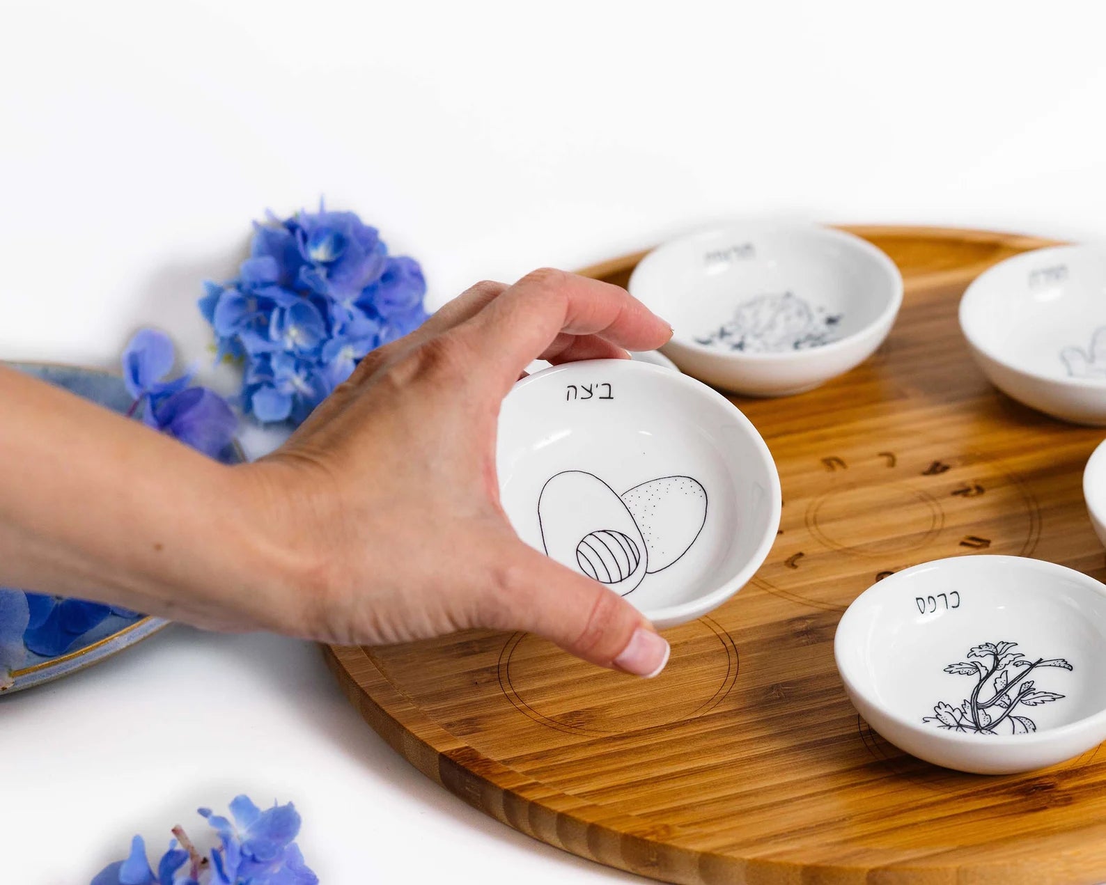 Closeup of Bamboo Passover Seder plate with porcelain bowls featuring black and white illustrations with a hand holding the egg bowl
