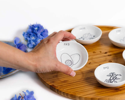 Closeup of Bamboo Passover Seder plate with porcelain bowls featuring black and white illustrations with a hand holding the egg bowl