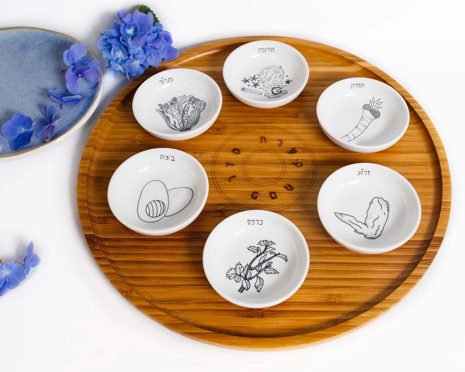 Closeup of Bamboo Passover Seder plate with porcelain bowls featuring black and white illustrations next to blue flowers