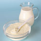 pitcher of milk filled to the top behind glass bowl of whey milk powder with blue background