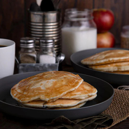 two seperate stacks of buttermilk pancakes on two black plates with salt coffee milk and apples in the background