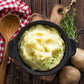 fluffy mashed potatoes in black dish on wooden cutting board with whole potatoes and herbs on the side and wooded spoon on plaid cloth
