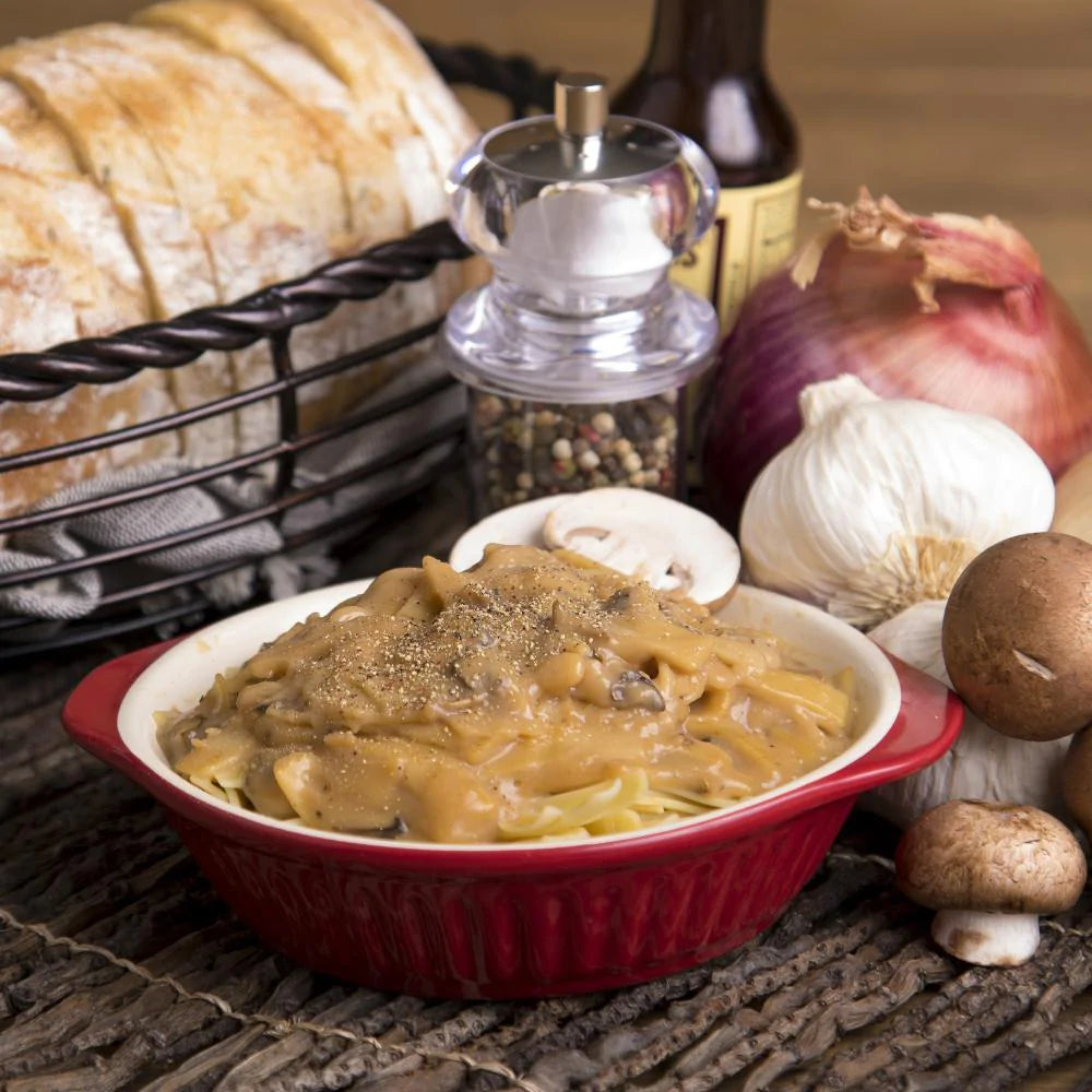 creamy stroganoff  in red dish surrounded by mushrooms red and white onions a loaf of bread and pepper 