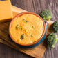 cheesy broccoli soup with extra shredded cheese on top in blue and yellow bowl on wooden cutting board with three heads of broccoli at side of bowl